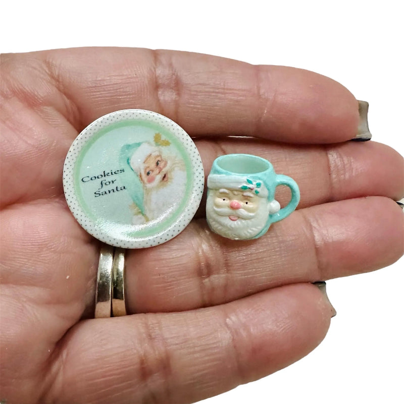 Two miniature Christmas-themed items on a hand, one resembling a cookie plate with Santa Claus and the other a teacup with a Santa face.