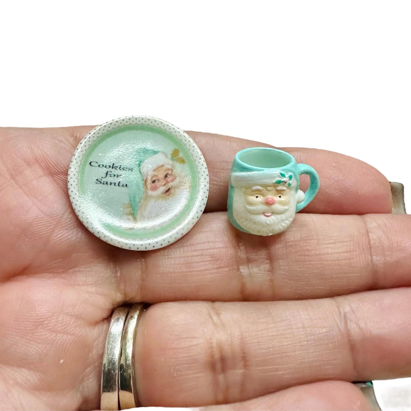 Two miniature Christmas-themed items on a hand, one resembling a cookie plate with Santa Claus and the other a teacup with a Santa face.