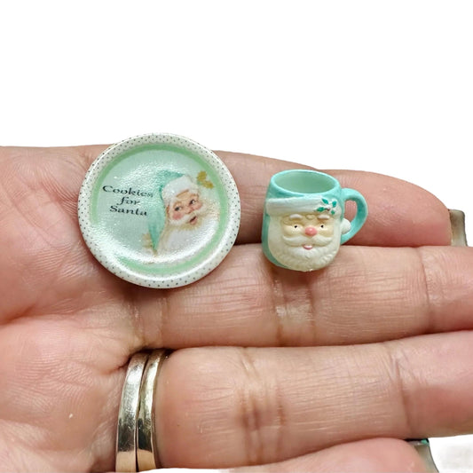 Two miniature Christmas-themed items on a hand, one resembling a cookie plate with Santa Claus and the other a teacup with a Santa face.